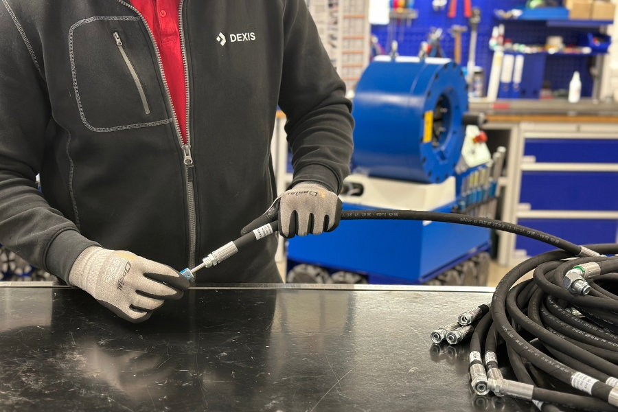 Ein Mann mit Handschuhen und dunkler Jacke arbeitet mit einem Schlauch auf einem schwarzen Tisch. Aufgerollte Schläuche mit Metallverbindern sind sichtbar, im Hintergrund ist eine blaue Hydraulikschlauchpresse zu sehen.